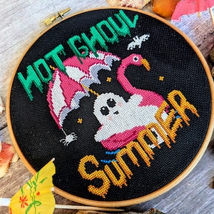 May include: A black embroidery hoop with a cross-stitch design of a ghost riding a pink flamingo with an umbrella. The text "Not ghoul summer" is stitched in green and yellow.
