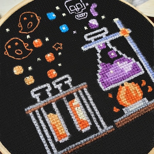 May include: A black embroidery hoop with a Halloween-themed design. The design features a purple potion bottle, orange and white ghosts, a skull, and orange and white stars. The design is stitched on black fabric.