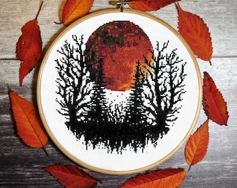 Puede incluir: Un aro de bordado de punto de cruz con un diseño en blanco y negro de árboles silueteados contra una luna roja y naranja. El aro está rodeado de hojas de otoño naranja y marrón.