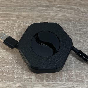 Könnte beinhalten: Ein schwarzer sechseckiger Kabelorganizer mit einem schwarzen Kabel, das darin aufgewickelt ist. Das Kabel hat an einem Ende einen USB-A-Stecker und am anderen Ende einen Lightning-Stecker.
