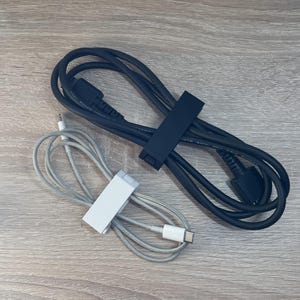 Könnte beinhalten: Zwei Kabel, eines schwarz und eines weiß, sind aufgerollt und mit Kabelbindern befestigt. Das schwarze Kabel hat einen größeren Stecker am Ende, während das weiße Kabel einen kleineren Stecker hat.
