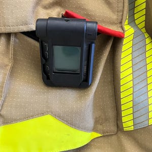 Peut inclure: Un appareil de sécurité incendie noir et rouge avec un écran numérique, porté dans une poche sur une veste ignifuge brune et jaune.
