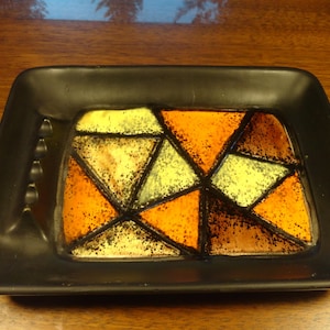 Op de afbeelding: Een zwarte keramische dienblad met een geometrisch ontwerp in tinten geel, oranje en bruin. Het ontwerp lijkt op glas-in-lood.
