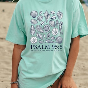May include: A light mint green short-sleeve t-shirt featuring a dark blue graphic print of various seashells and sand dollars. Below the shells, the text reads PSALM 95:5 THE SEA IS HIS, FOR HE MADE IT in a dark blue sans-serif font.