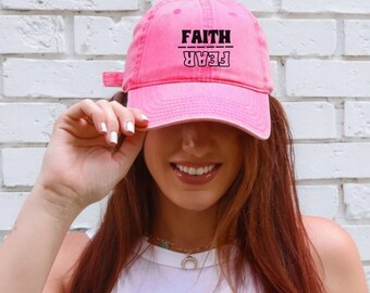 Puede incluir: Gorra de béisbol rosa con las palabras "FAITH" sobre "FEAR" en negro. La gorra la lleva una persona, con la visera proyectando una sombra. El fondo es una pared de ladrillos blancos.