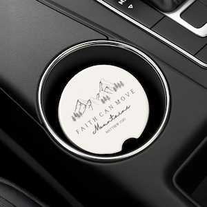 May include: A white car coaster with a black mountain and tree design. The coaster has the text "FAITH CAN MOVE Mountains" and "Matthew 17:20" in black script. The coaster is in a car cup holder.