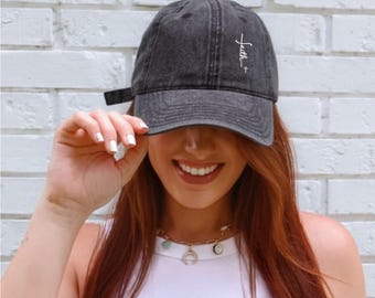 May include: A dark gray baseball cap with the word "faith" and a cross embroidered in white. The cap is worn by a person with long red hair, a white top, and a gold necklace. The background is a white brick wall.