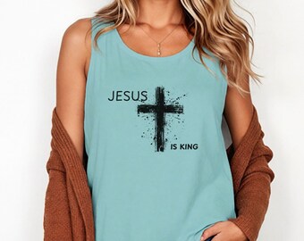 May include: A light blue tank top with the words "JESUS IS KING" printed in black. The design includes a black cross with a splatter effect. The top is sleeveless and has a scoop neck.