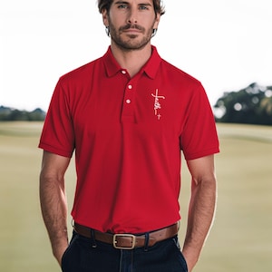 May include: A red polo shirt with short sleeves and a collar. The shirt has a white "Faith +" script design on the left chest. The model is wearing a brown belt and blue jeans. The background is a green field.