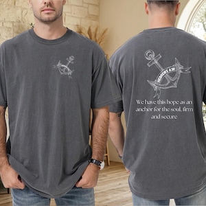 Op de afbeelding: Donkergrijs t-shirt met een witte ankerprint op de voor- en achterkant. De achterkant van het shirt heeft de tekst "HEBREWS 6:39" en een citaat over hoop. Het t-shirt is gemaakt van een zacht, comfortabel materiaal.
