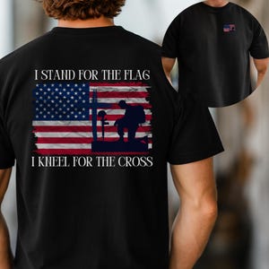 Könnte beinhalten: Schwarzes T-Shirt mit einer Grafik der amerikanischen Flagge, einem Kreuz und einem knienden Soldaten. Der Text lautet "I STAND FOR THE FLAG" und "I KNEEL FOR THE CROSS". Eine kleine amerikanische Flagge befindet sich auf der oberen linken Brust.