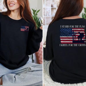 May include: Black long-sleeve sweatshirt featuring an American flag design with a soldier and cross graphic. The front has a small flag graphic. The back displays the text "I STAND FOR THE FLAG, I KNEEL FOR THE CROSS."