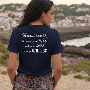 Pode incluir: Camiseta azul marinho com texto branco: "Accept what IS, let go of what WAS, and have faith in what WILL BE." A pessoa usa a camiseta ao ar livre, com um fundo costeiro de mar e edifícios.
