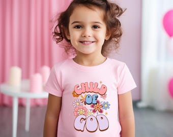 Puede incluir: Una camiseta rosa claro con las palabras "CHILD OF GOD" en una fuente retro, decorada con flores coloridas. La camiseta la lleva una niña pequeña con el pelo rizado castaño.