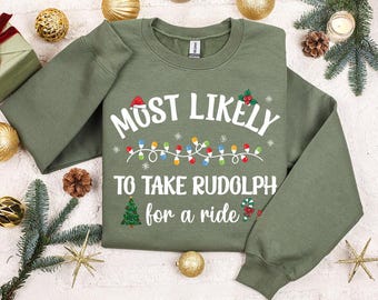 May include: An olive green sweatshirt with the text "MOST LIKELY TO TAKE RUDOLPH for a ride" in white. The design includes a Santa hat, Christmas lights, snowflakes, a Christmas tree, and a candy cane. Festive holiday apparel.
