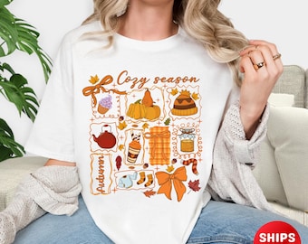 May include: White t-shirt with a colorful autumn-themed graphic. The design includes pumpkins, a teapot, a cupcake, a jar of honey, and the words "Cozy season" and "Autumn". The shirt has a crew neck and short sleeves.