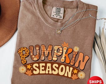 Op de afbeelding: Een bruin t-shirt met de tekst "PUMPKIN SEASON" in een retro lettertype. De letters zijn versierd met luipaardprint, bloempatronen en houtnerf. Een zilveren ketting met een hanger is zichtbaar. Het shirt maakt deel uit van een herfstcollectie.