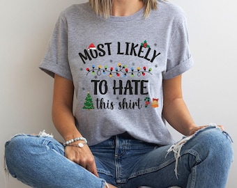 May include: A light gray t-shirt with the text "MOST LIKELY TO HATE this shirt" in black, red, green, and white. The design includes a Santa hat, Christmas lights, a Christmas tree, and a gingerbread house.