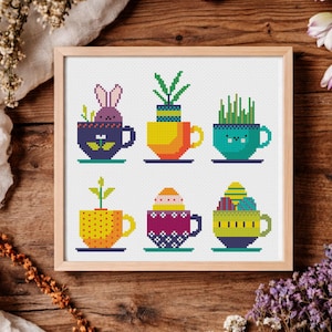 Puede incluir: Un bordado de punto de cruz enmarcado con seis tazas de té coloridas, cada una con un diseño único. Las tazas contienen un conejito, plantas y huevos de Pascua. La paleta de colores incluye azul, amarillo, naranja y verde. La obra está sobre una superficie de madera.