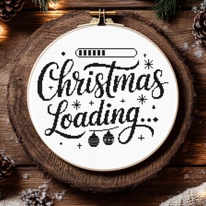 Puede incluir: Un bordado en punto de cruz blanco con la frase "Christmas Loading..." en negro. El diseño incluye una barra de carga, copos de nieve y adornos. El bastidor está colocado sobre una rodaja de madera.