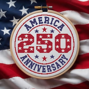 Op de afbeelding: Een borduurring met kruissteek met de tekst "AMERICA 250 ANNIVERSARY" in rood en blauw, met sterren. De ring staat tegen een achtergrond van de Amerikaanse vlag, met rode en witte strepen en een blauw veld met sterren.