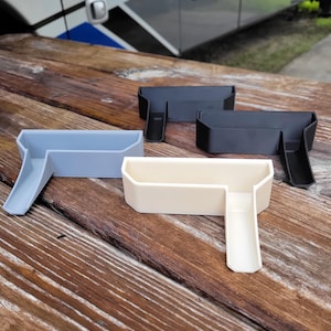 May include: Four plastic RV accessories in light blue, black, and cream colors. Each has a rectangular tray with a ramp-like extension. The objects are arranged on a wooden surface, with a recreational vehicle visible in the background.