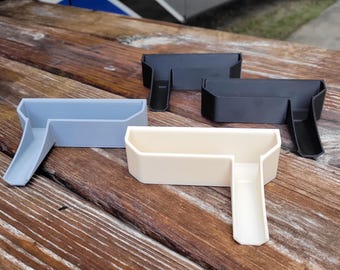 May include: Four plastic RV accessories in light blue, black, and cream colors. Each has a rectangular tray with a ramp-like extension. The objects are arranged on a wooden surface, with a recreational vehicle visible in the background.