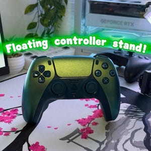 Op de afbeelding: Een gamecontroller met een metallic groene en blauwe kleurveranderende afwerking, voorzien van standaard richtingsknoppen, twee analoge sticks en het PlayStation-logo. De tekst "Floating controller stand!" staat in het groen boven het apparaat.