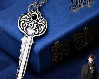 May include: A silver key necklace with a detailed design and the number "221B" engraved on it. The necklace is on a blue velvet background with the words "Sherlock Holmes" in a decorative font.