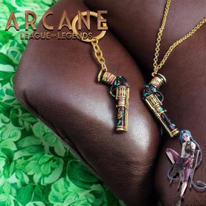 Op de afbeelding: Een goudkleurige sleutelhanger en ketting met een pistoolontwerp met zwarte, gouden, teal en roze accenten. De sleutelhanger heeft de tekst "ARCANE LEAGUE OF LEGENDS". Een Jinx-personage-uitsparing is ook zichtbaar.