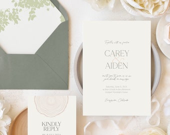 May include: Wedding invitation suite with a green envelope, a white invitation card with the text "Carey & Aiden" and a white RSVP card with a wood grain design. The invitation suite is surrounded by white and peach flowers.