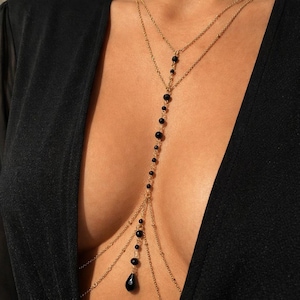 May include: A gold-toned body chain necklace featuring two delicate upper chains and a central vertical strand adorned with small black beads. The design culminates in a single black teardrop-shaped pendant, creating a layered look over a black garment.