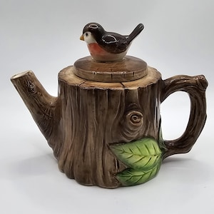 May include: A ceramic teapot in the shape of a tree stump, with a brown and green colour scheme. The teapot has a handle, spout, and a lid topped with a small bird figurine. Green leaves are painted on the side.
