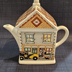 May include: A ceramic teapot shaped like a two-story house with a brown roof. The front features illustrations of people in windows, a yellow and black car labelled "INGTONS TEA," and figures standing outside a brick building with a green door.