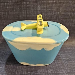 May include: A light blue and white ceramic butter dish with a cloud-like pattern. The lid features a yellow biplane figurine on top with the text Utterly Butterly printed in black on the wing, serving as a handle for the oval-shaped container.