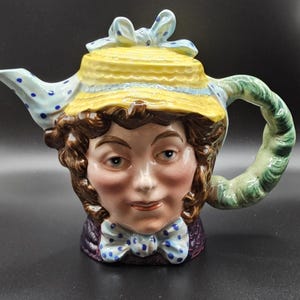 May include: A ceramic teapot in the shape of a woman's face, adorned with a yellow hat and blue bow. The spout is white with blue spots, and the handle is green. The teapot features brown hair, a purple jacket, and a blue polka-dot bow tie.