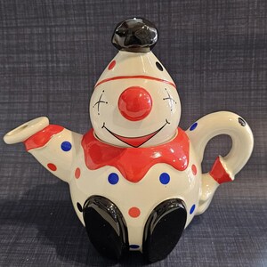 May include: A ceramic teapot shaped like a clown with a cream-coloured body, a red nose, and a black hat. The teapot features a red collar, black shoes, and a pattern of red and blue polka dots across the surface. It has a curved handle and a spout.