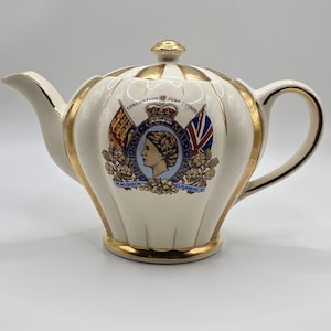 May include: A cream-coloured ceramic teapot with gold trim. It features a central emblem with a profile portrait and the text "Coronation June 2 1953." Includes flags and decorative elements, a spout, handle, and lid.