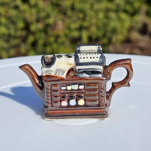 May include: A brown ceramic teapot in the form of a vintage cash register. The teapot has a spout, handle, and a detailed design with various colours, including white, black, and pastel shades. The teapot is set against a blurred green background.