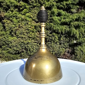 May include: A vintage brass bell with a black wooden handle. The bell has a rounded base and a long, slender stem. The handle is a dark, ribbed design. The bell is sitting on a white surface with a green background.