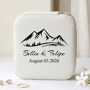 Könnte beinhalten: Eine weiße, quadratische Schmuckschatulle mit einem schwarzen Bergketten-Design und den Namen "Billie & Felipe" mit dem Datum "August 03.2026" darunter. Die Box hat einen Reißverschluss und steht auf einer weißen Oberfläche.