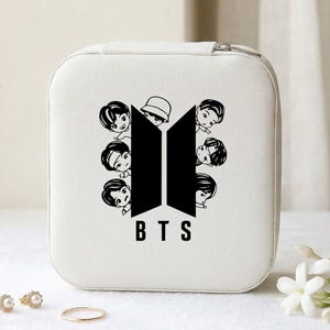 May include: A white, square jewelry box with a black graphic of the BTS logo and cartoon figures. The letters "BTS" are printed in black below the logo. A gold ring and two earrings are in the foreground.