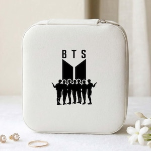 May include: A white, square jewelry box with a zipper closure. The front features a black graphic of the letters "BTS" above a silhouette of seven figures. Gold rings and earrings are in the foreground.