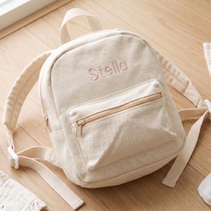 Könnte beinhalten: Beigefarbener Canvas-Rucksack mit dem Namen "Stella" in Rosa gestickt. Der Rucksack hat eine Reißverschlusstasche vorne, verstellbare Träger und einen oberen Griff. Babyartikel, darunter Socken und eine Decke, sind ebenfalls sichtbar.