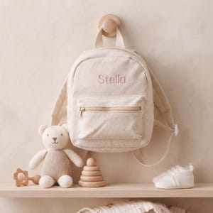 Könnte beinhalten: Ein cremefarbener Rucksack aus Segeltuch mit dem Namen Stella, der in hellrosa Garn auf die Vordertasche gestickt ist. Der Rucksack verfügt über einen Tragegriff, verstellbare Schultergurte und ein Reißverschlussfach vorne und hängt an einem Holzhaken.