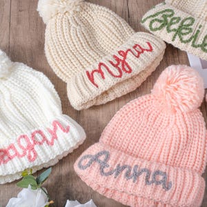 Könnte beinhalten: Eine Auswahl an Strickmützen in Creme, Rosa und Weiß, jeweils mit Bommel und einem Namen, der auf den Rand gestickt ist. Die Namen sind in Schreibschrift gehalten. Die Mützen sind auf einer Holzoberfläche angeordnet.