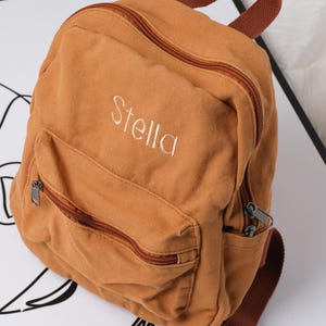 May include: A tan canvas backpack with a front zipped pocket and a top handle. The name "Stella" is embroidered in white on the front. The backpack has brown zips and straps.