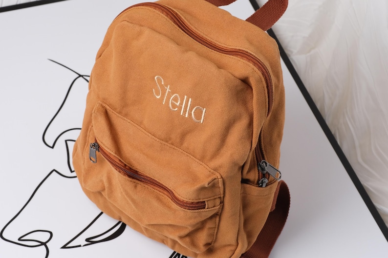 May include: A tan canvas backpack with a front zipped pocket and a top handle. The name "Stella" is embroidered in white on the front. The backpack has brown zips and straps.