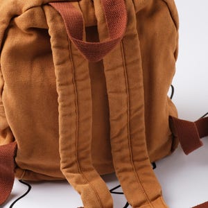 May include: A close-up of a brown canvas backpack with adjustable shoulder straps. The backpack has a zippered pocket and a top handle. The straps and handle are a darker shade of brown.