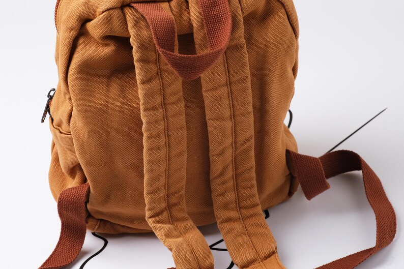 May include: A close-up of a brown canvas backpack with adjustable shoulder straps. The backpack has a zippered pocket and a top handle. The straps and handle are a darker shade of brown.